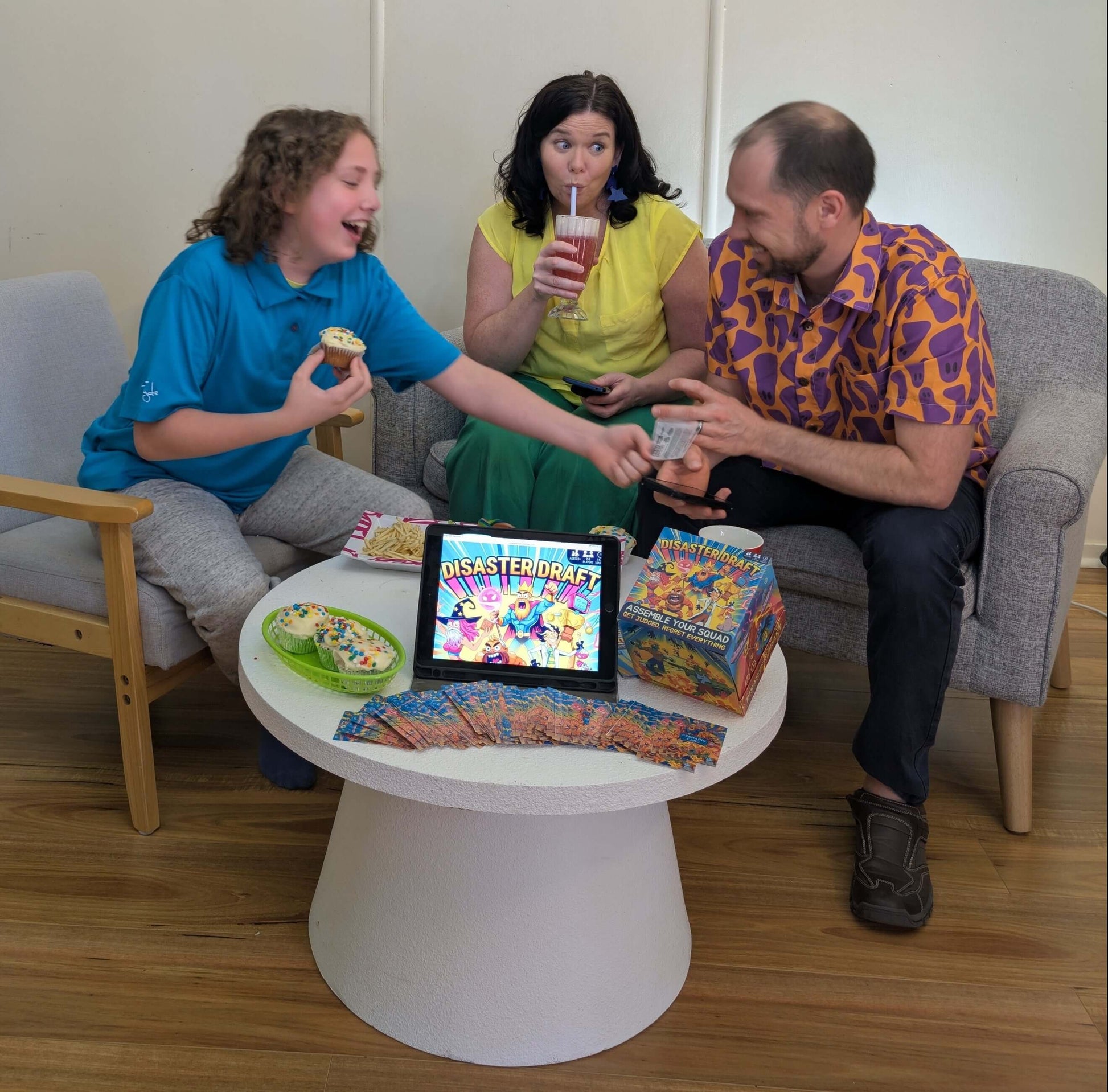 Three people sitting around a small table with a tablet displaying the Companion App game of Disaster Draft ,and the cards on the table. They are having a lot of fun playing the game