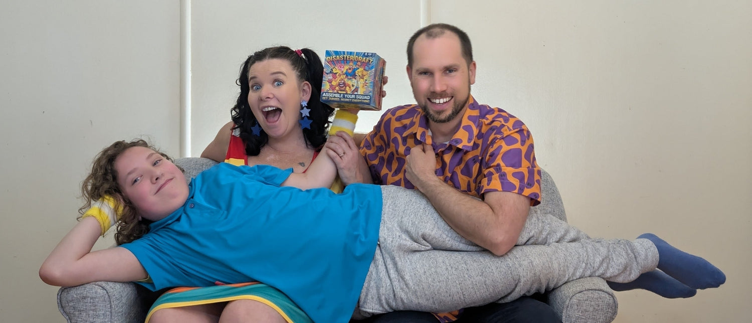 Two people sitting on a couch with a child on their lap in a room with a white wall. They are holding a copy of the Australian Card Game Disaster Draft
