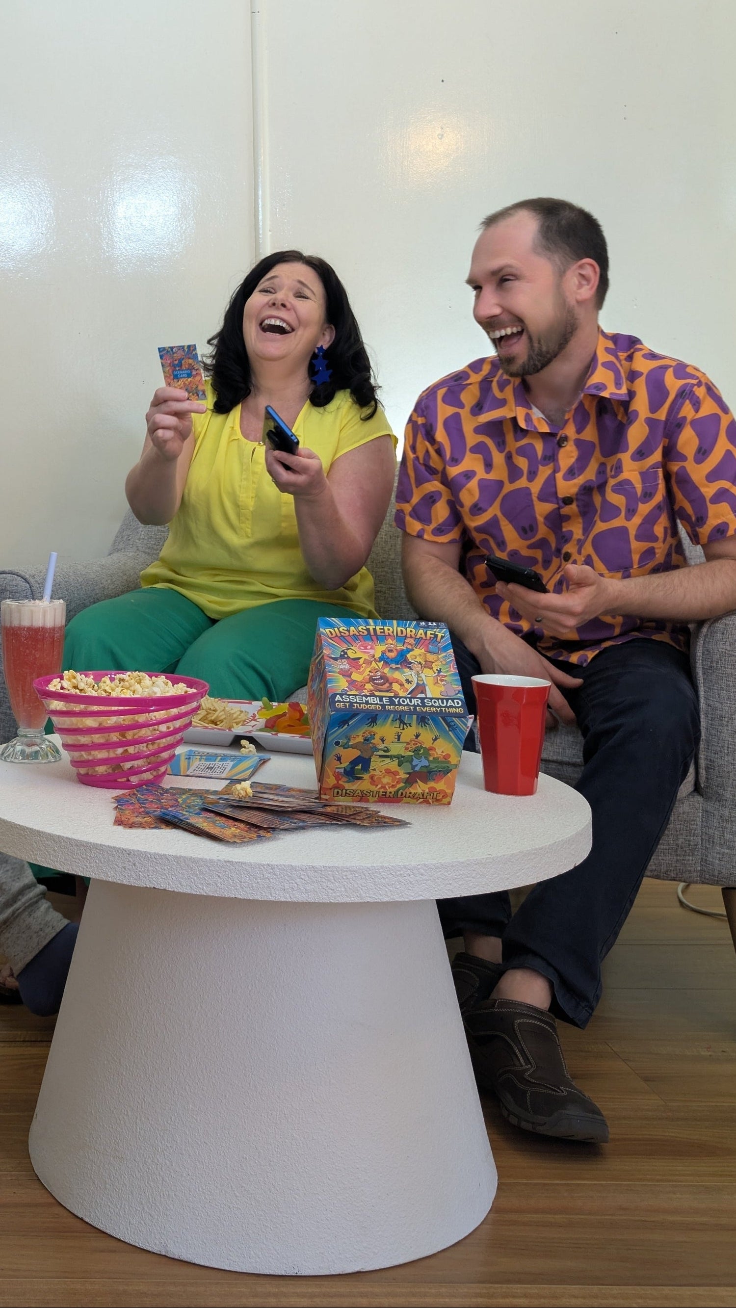 Two adults are laughing and playing disaster draft. They are sitting together at a table with snacks and drinks. They game box is visible in the front